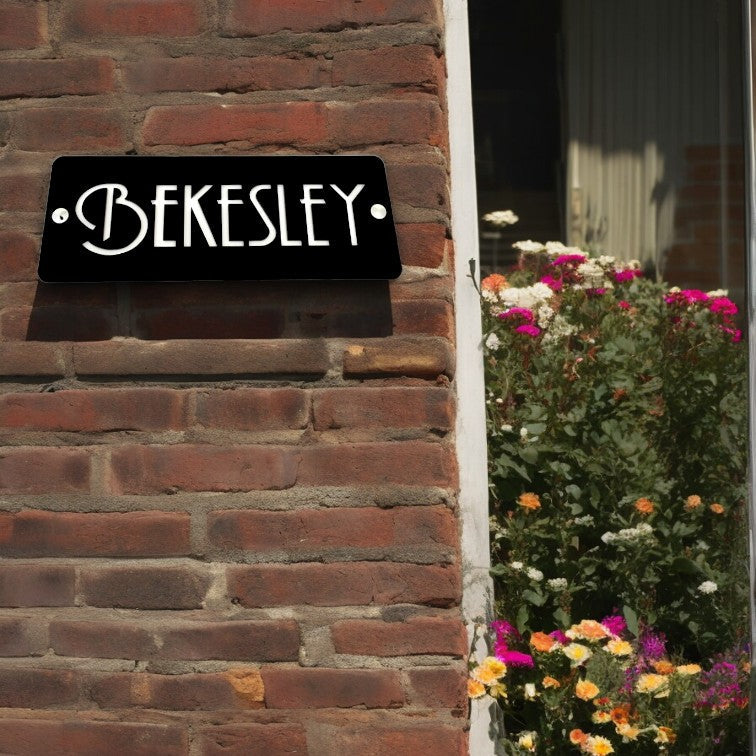 Rectangular black house sign with white "BEKESLEY" text in art deco style, mounted on a red brick wall.