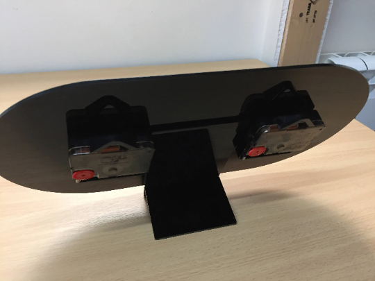 Back view of a black oval desk clock stand with two clock mechanisms attached, resting on a wooden surface.