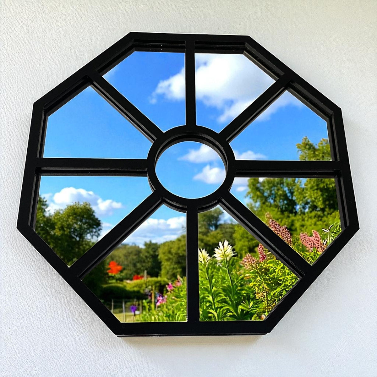 An octagonal black garden mirror with paneled muntins reflecting a garden, trees, and sky.