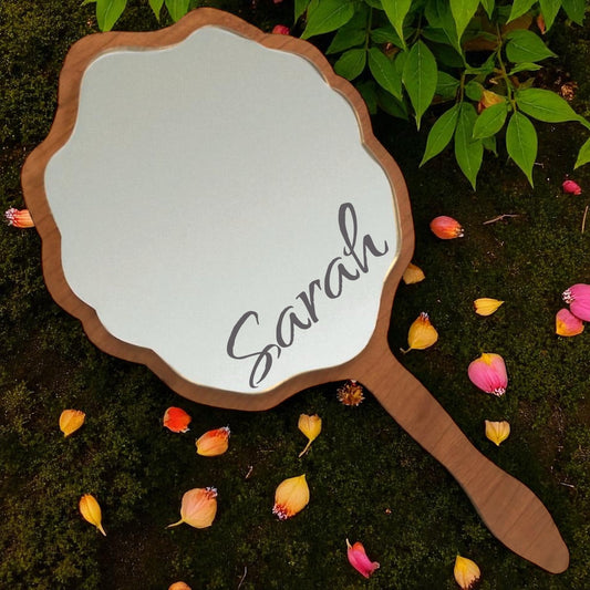 Scalloped wooden hand mirror with the engraved name Sarah, resting on moss among fallen petals.