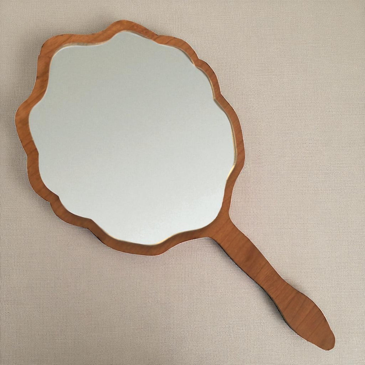 Scalloped hand-held wooden vanity mirror with a light brown frame and handle on a textured white background.