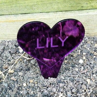 Personalised purple heart-shaped mirror memorial sign with the name Lily engraved, planted in soil.