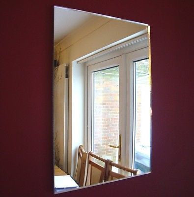 Frameless rectangular mirror hanging on a dark red wall, reflecting an interior room with a door and chairs.