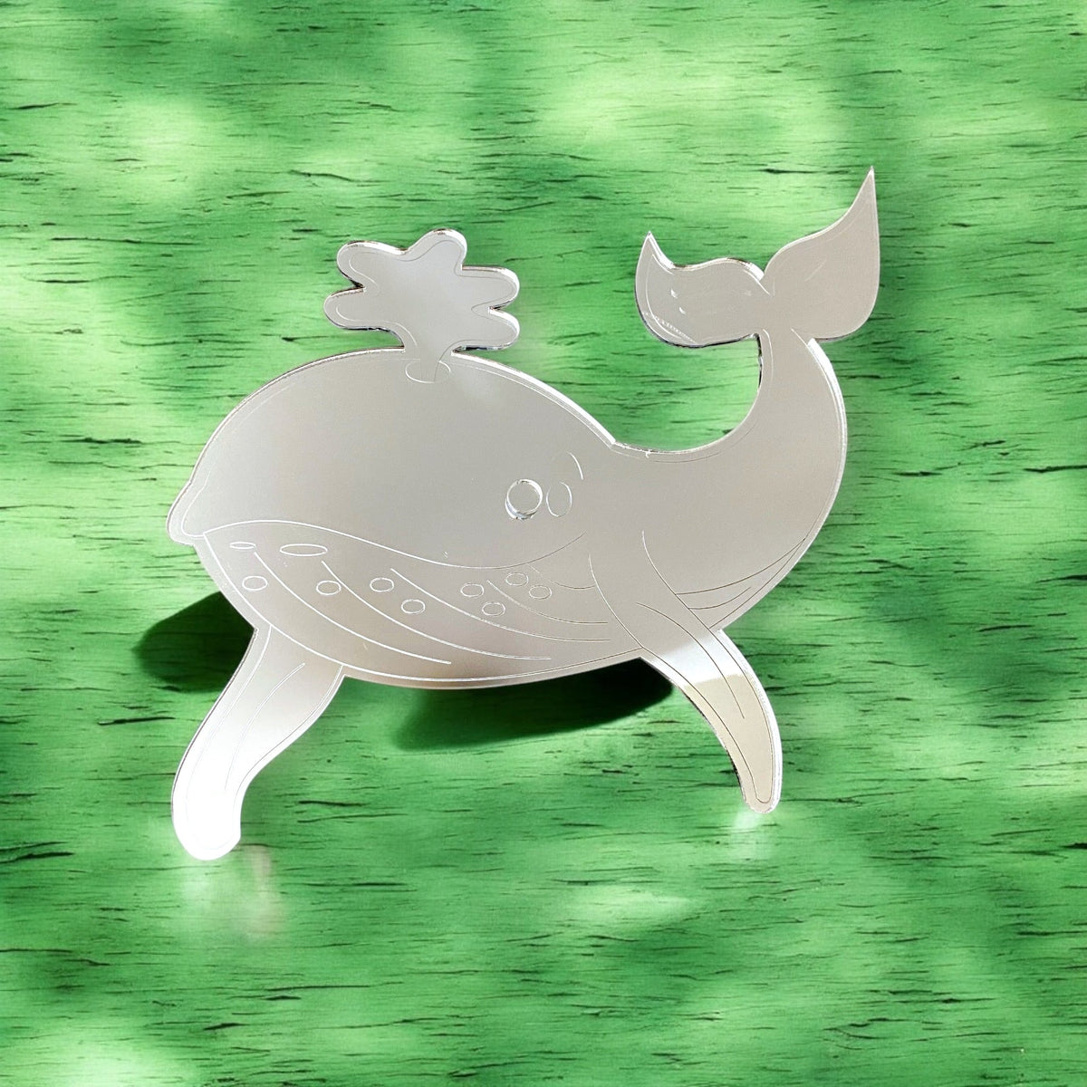 Etched whale-shaped mirror with water spout detail on a green wooden background.