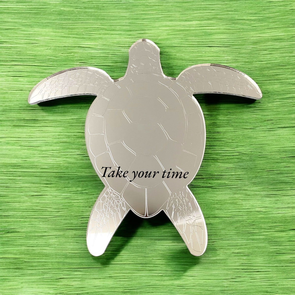 An etched silver turtle-shaped mirror with the text "Take your time" on a green surface.