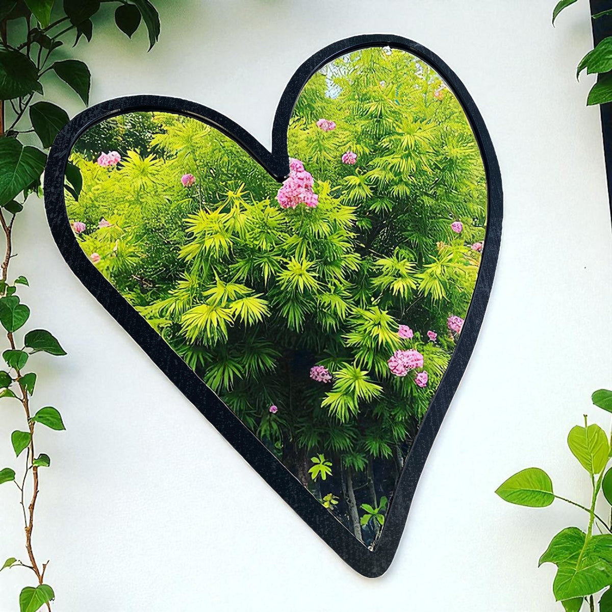 Black heart-shaped garden mirror hanging on a white wall, reflecting green plants.