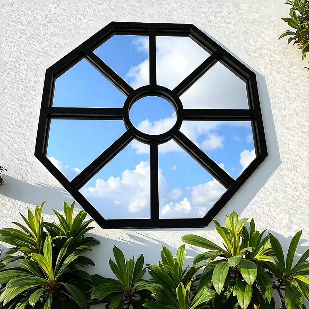 Octagonal black metal garden mirror mounted on a white wall, reflecting a cloudy blue sky.