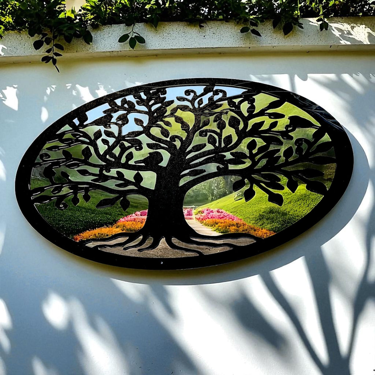 Oval black metal tree of life garden mirror hanging on a white wall, reflecting a green landscape.