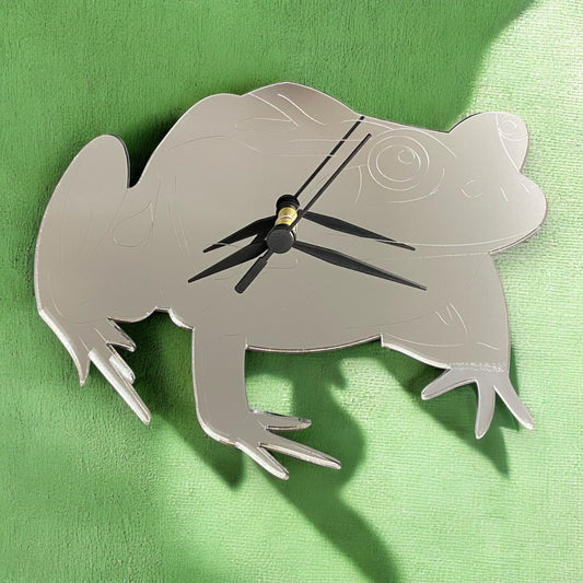 A silver mirror-finish, frog-shaped wall clock with black clock hands, mounted on a green textured background.