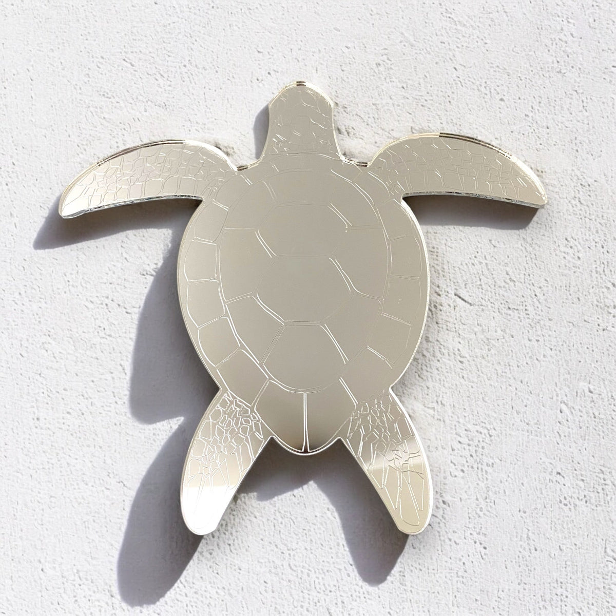 An etched silver turtle-shaped mirror mounted on a white textured wall.