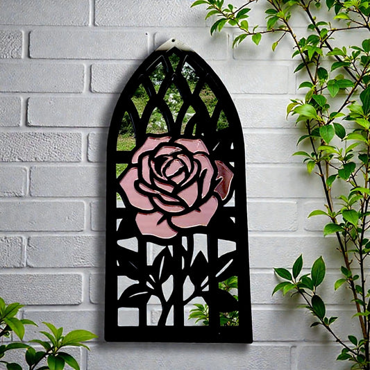Gothic church arch-shaped garden mirror with a black frame and a decorative pink rose inlay on a white brick wall.