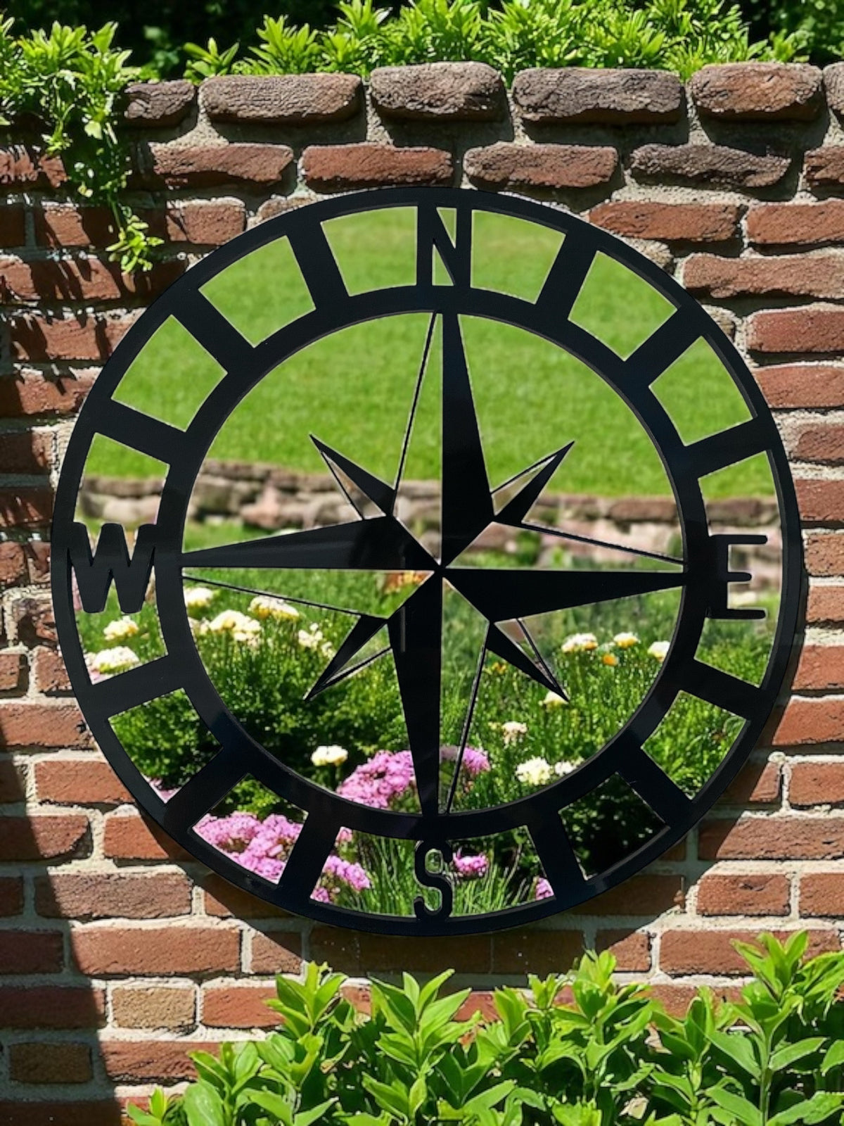 Black metal compass garden mirror mounted on a brick wall, reflecting a garden.