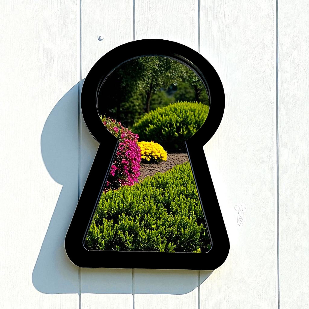 A black keyhole-shaped framed garden mirror hanging on a white wall, reflecting a garden.