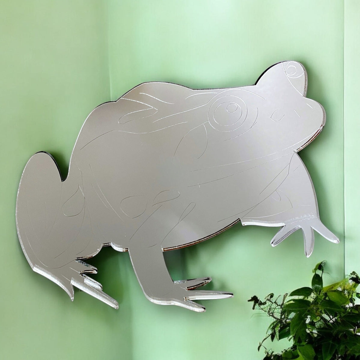 An etched frog-shaped mirror hangs on a light green wall above a small green plant.