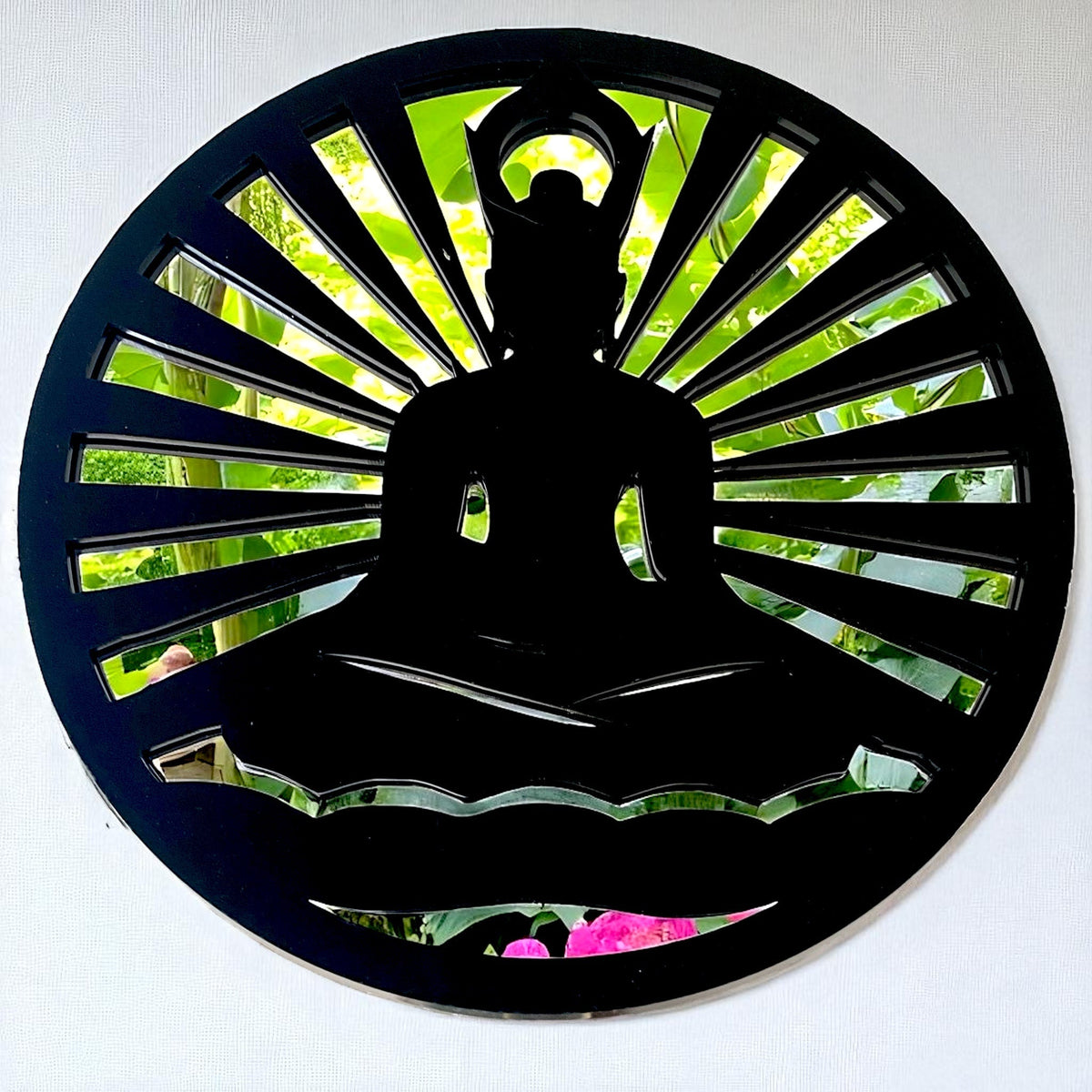 A round black mirror with a cutout silhouette of a meditating Buddha, reflecting green foliage.