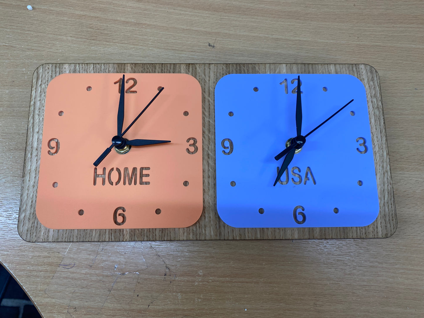 Two square clocks (orange and blue) on a wooden desk stand labeled "HOME" and "USA" with black hands.
