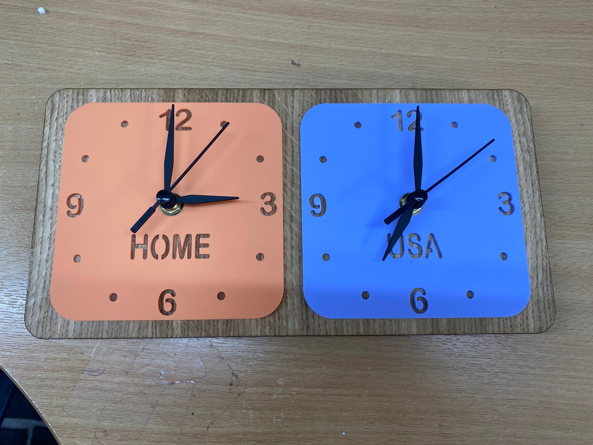 Two square clocks (orange and blue) on a wooden desk stand labeled "HOME" and "USA" with black hands.