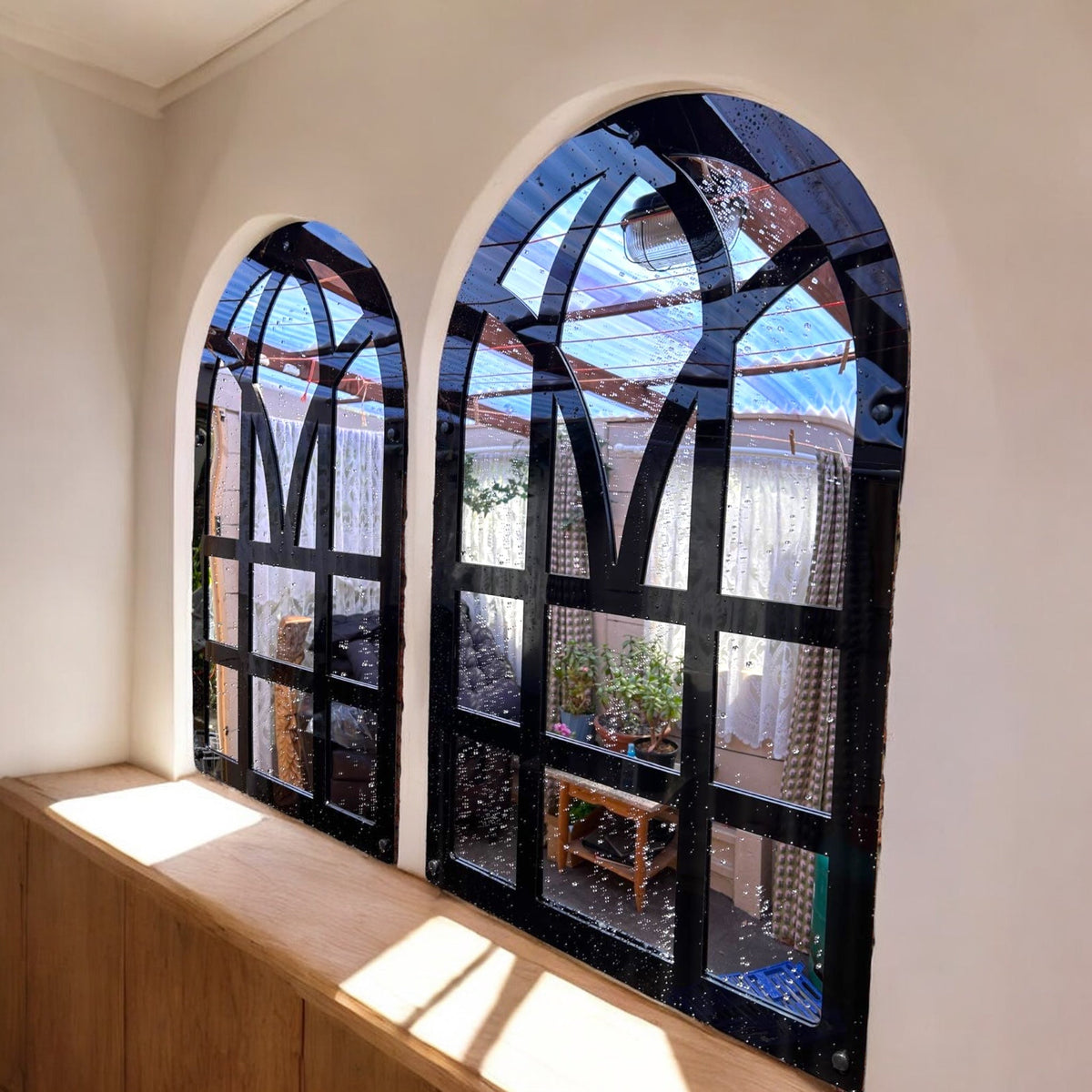 Pair of black Victorian arch garden mirrors with multi-pane frames, mounted on a white wall above a wooden ledge.