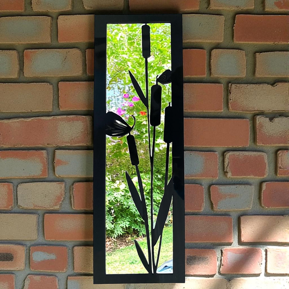 A vertical rectangular black framed garden mirror mounted on a red brick wall, with black metal bulrush silhouettes.