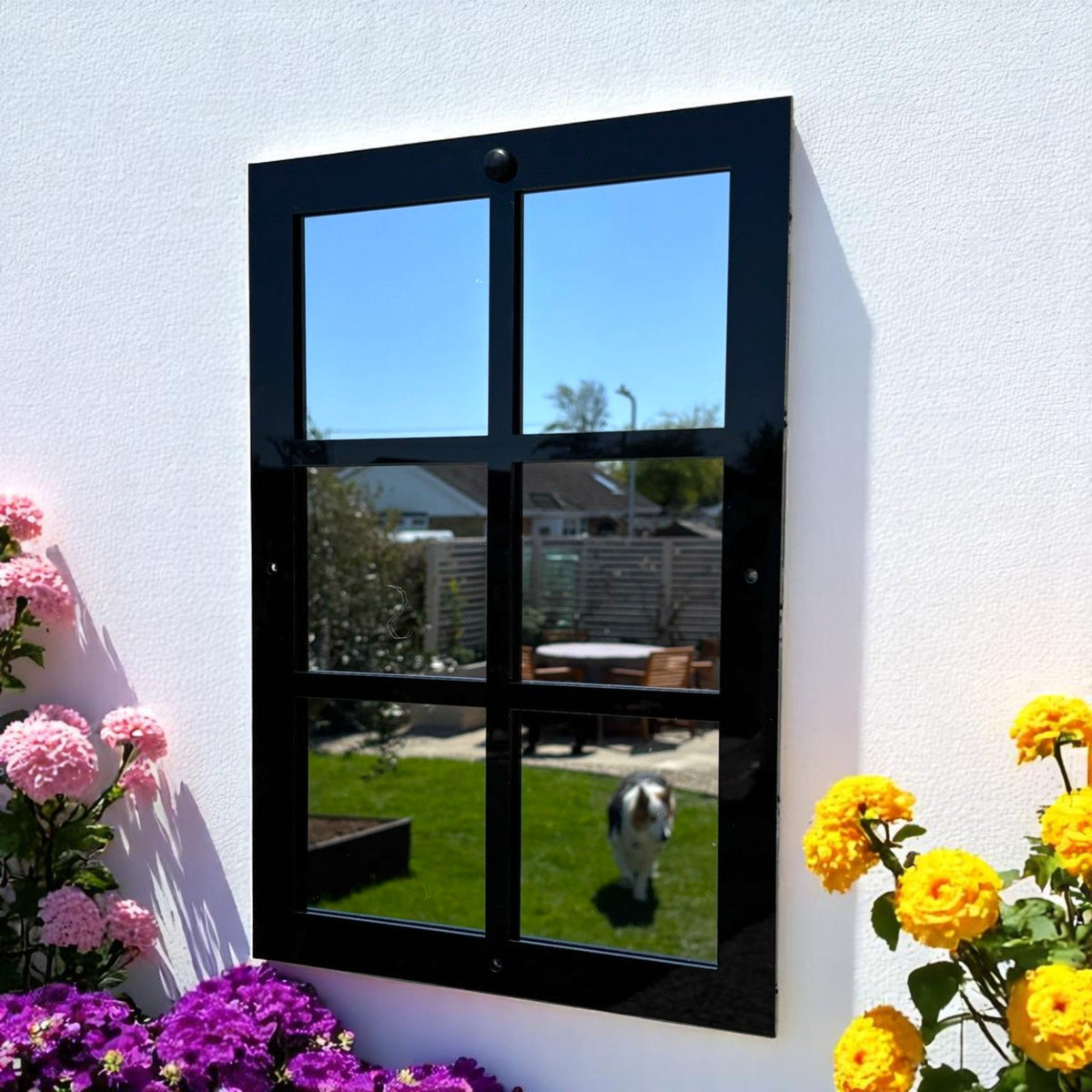 Six-panel black framed garden mirror mounted on a white wall, surrounded by flowers.