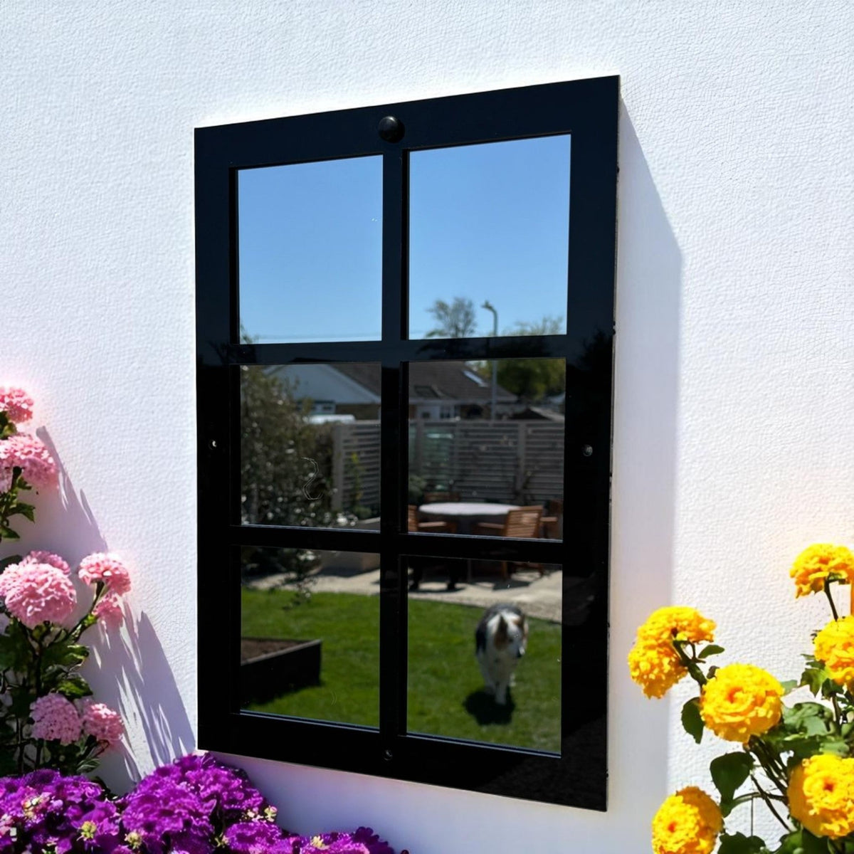 A black six-panel garden mirror mounted on a white wall, flanked by pink and yellow flowers.