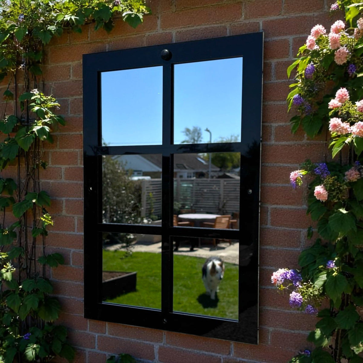 Black six-panel garden mirror mounted on a red brick wall, reflecting a sunny garden scene.