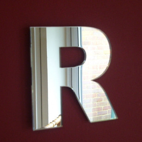 A reflective letter R mirror mounted on a dark red wall, reflecting a brick building and window.
