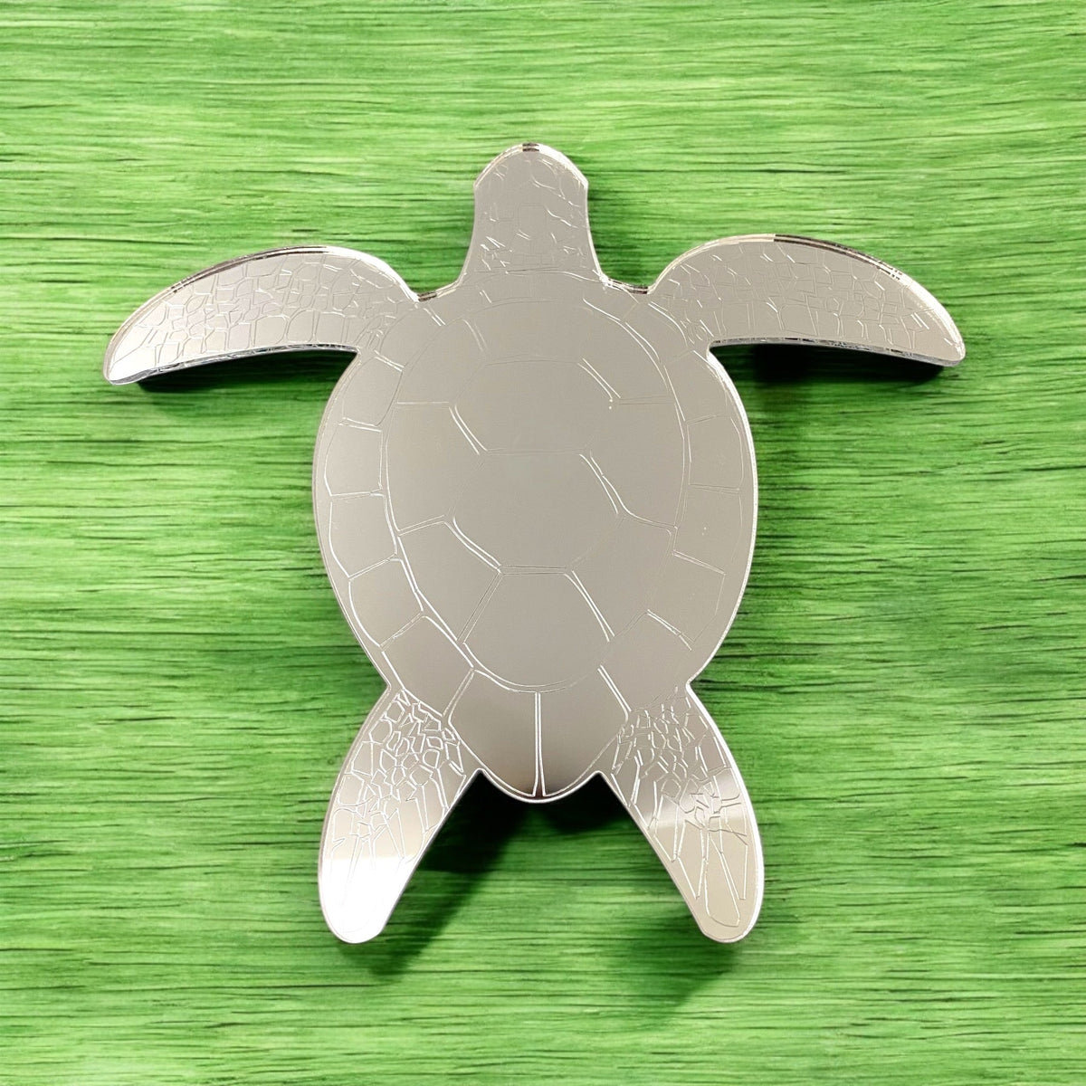 Silver turtle-shaped mirror with etched shell and flipper details, resting on a textured green surface.