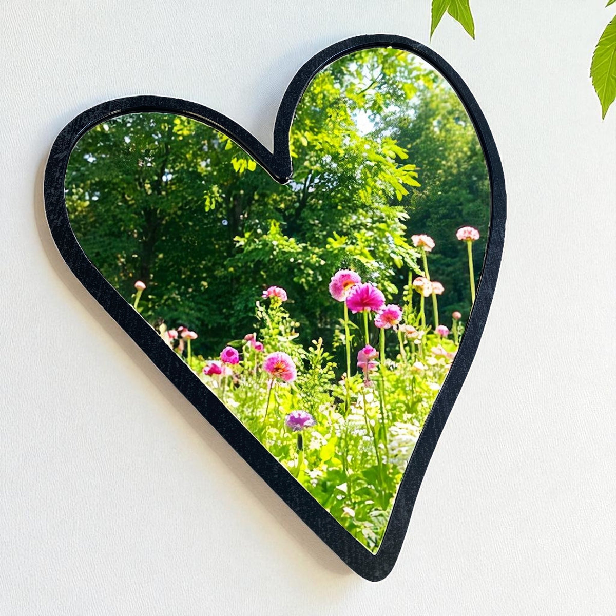 A heart-shaped mirror with a black frame, hanging on a wall, reflecting a garden with pink flowers and green trees.