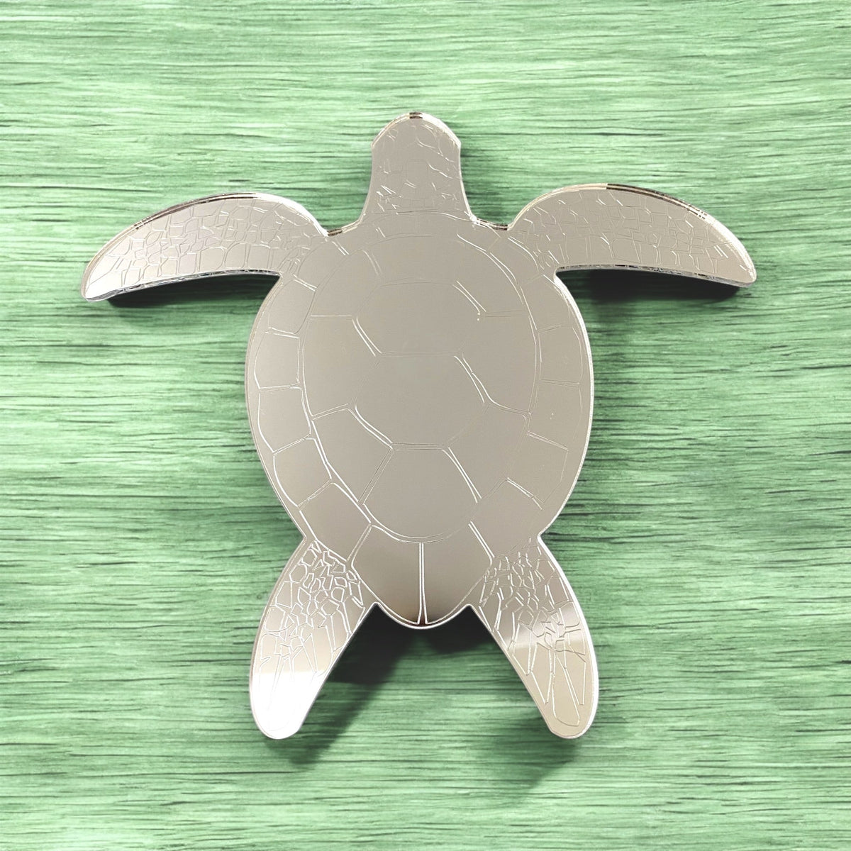 Etched silver sea turtle-shaped mirror on a textured green surface.
