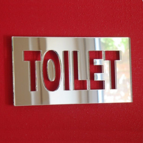 Rectangular mirrored toilet sign with cutout letters on a red surface.