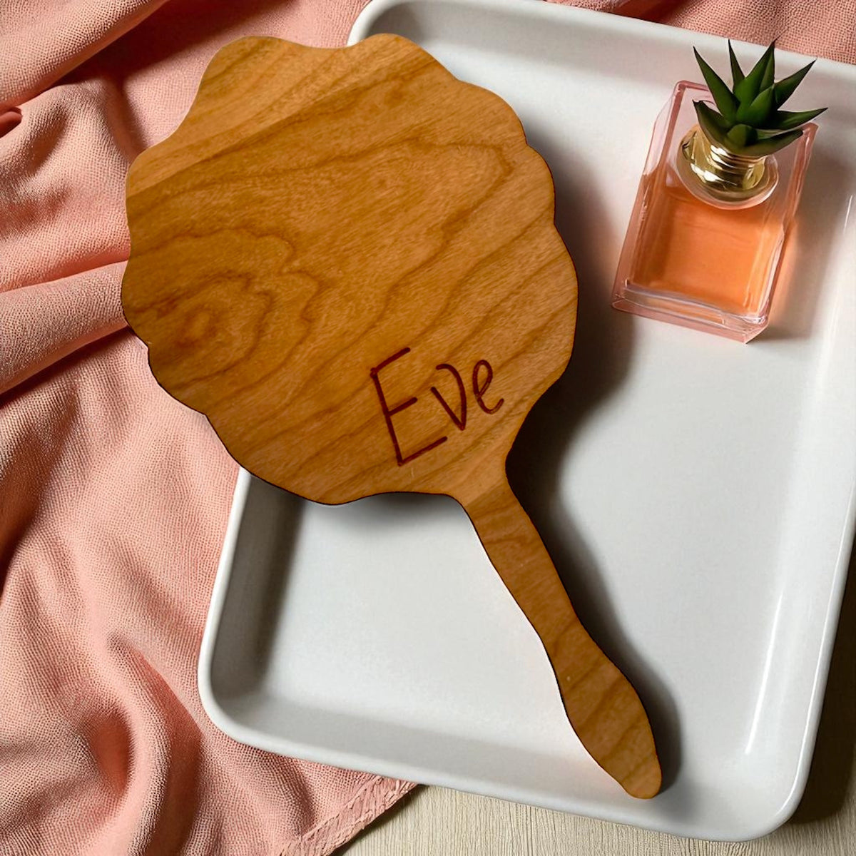 Scalloped wooden hand mirror with the engraved name Eve on a white tray, next to a pink perfume bottle.