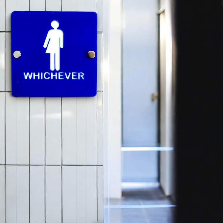 Square blue "WHICHEVER" toilet sign mounted on white tiled wall with silver standoffs.