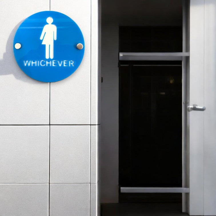Round blue acrylic toilet sign reading WHICHEVER with a white symbol, mounted on a tiled wall.