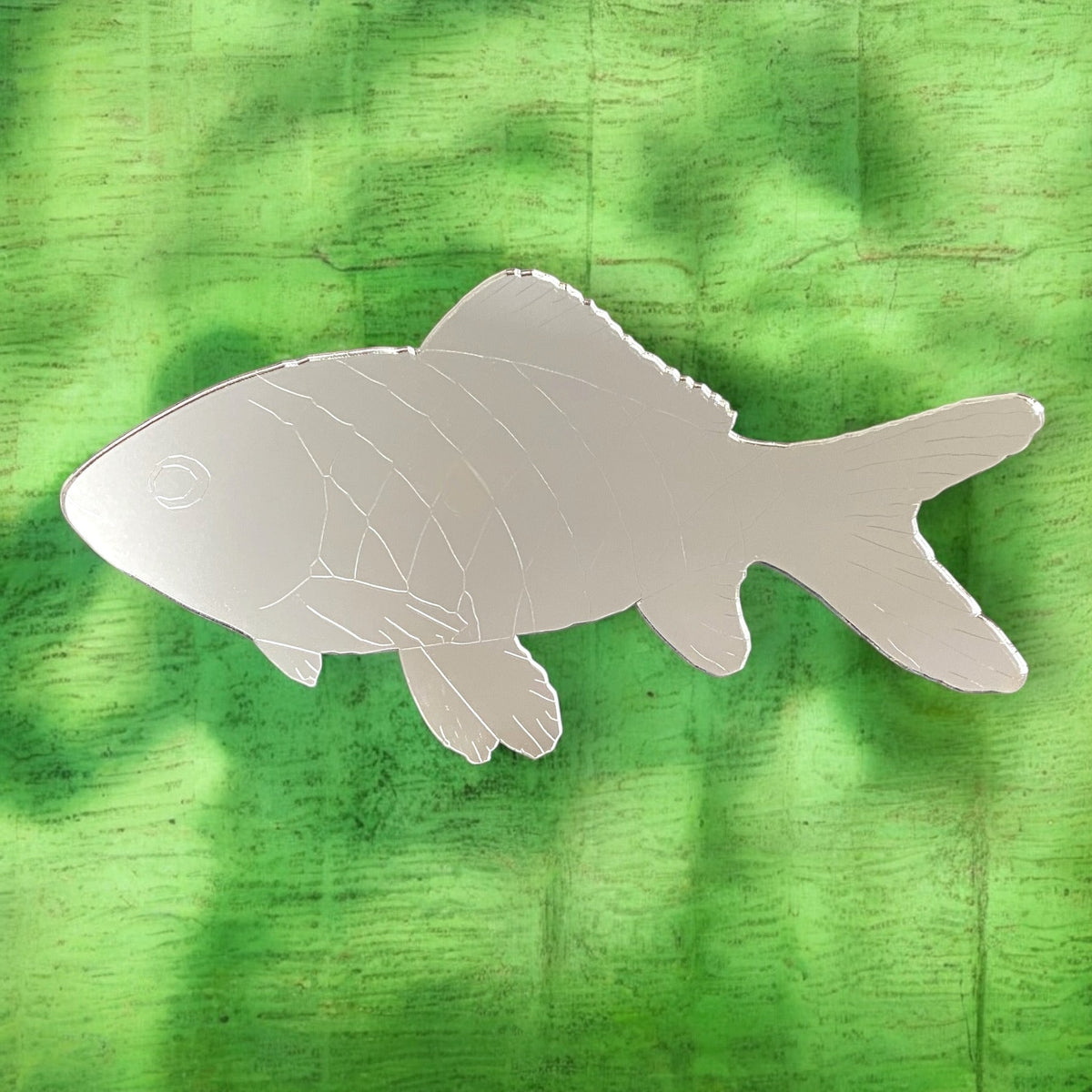 Silver fish-shaped mirror with etched scale lines, displayed against a textured green background.