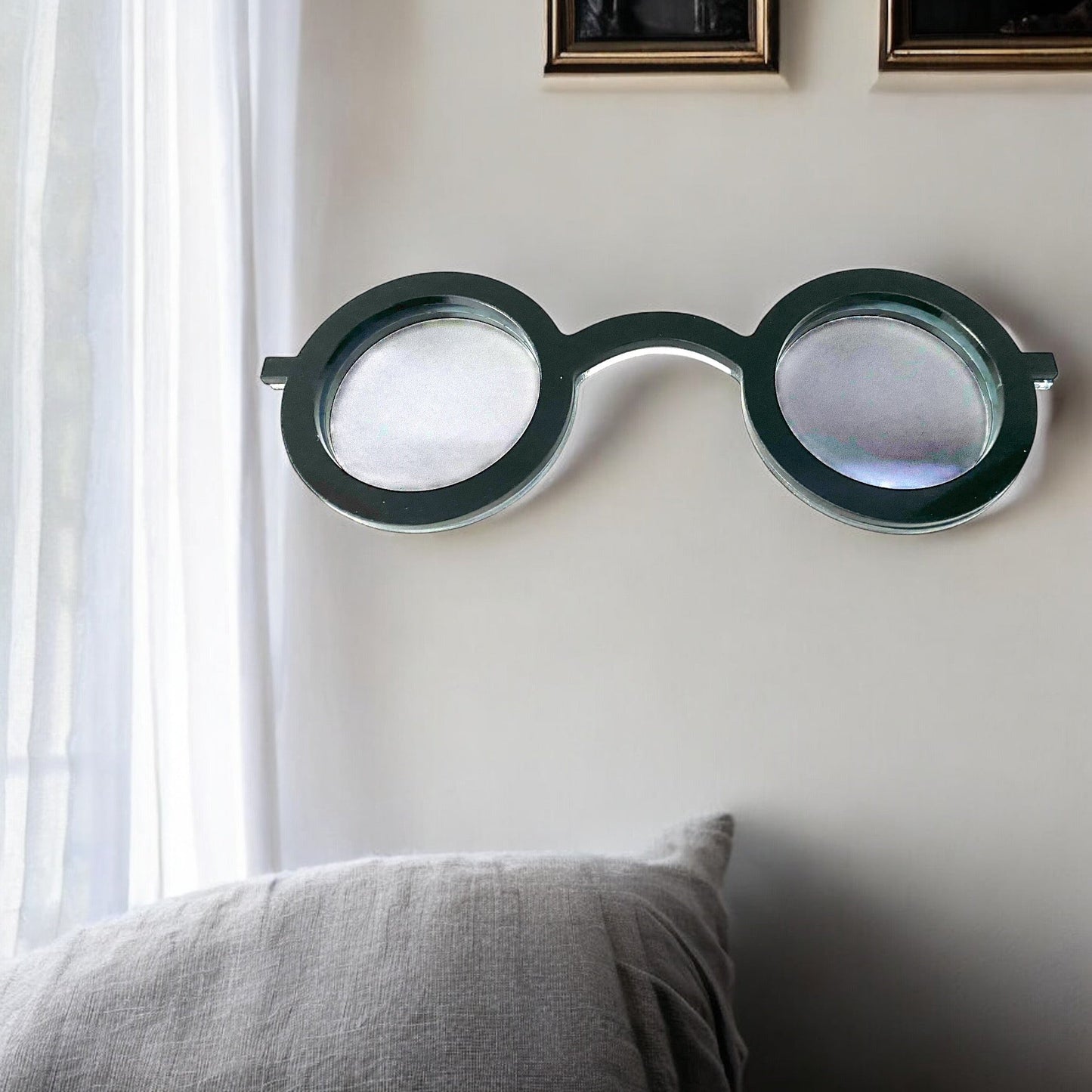 Round glasses-shaped wall mirror with a dark frame mounted on a white wall above a gray pillow.