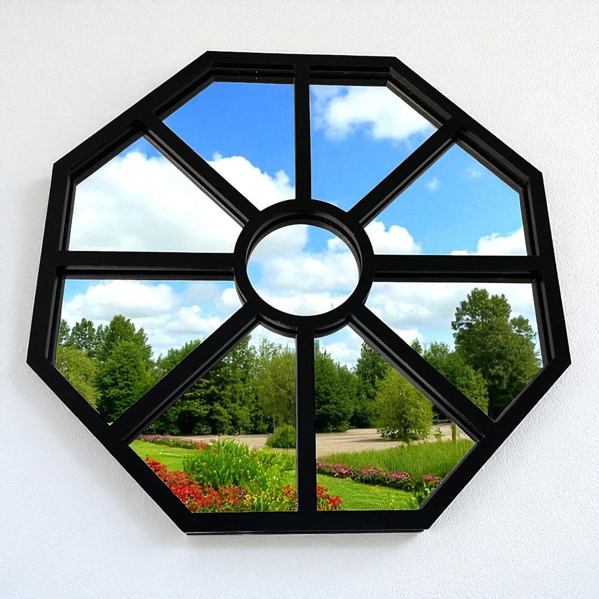 Octagonal black framed garden mirror hanging on a white wall, reflecting trees, grass, and a blue sky.
