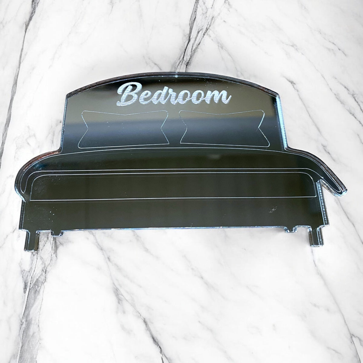 Engraved bed-shaped mirror on a white marble surface, featuring the word "Bedroom" and etched pillow designs.