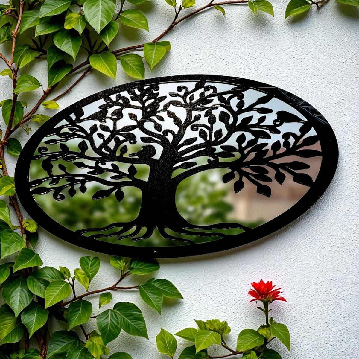 An oval garden mirror featuring a black metal Tree of Life overlay, mounted on a white wall amidst green foliage.