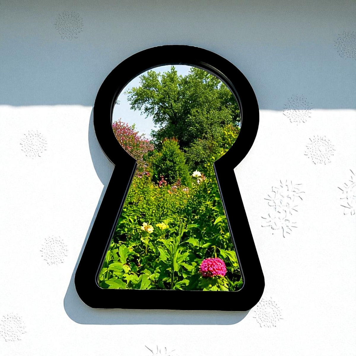 Keyhole-shaped black garden mirror mounted on a white wall, reflecting green trees and colorful flowers.