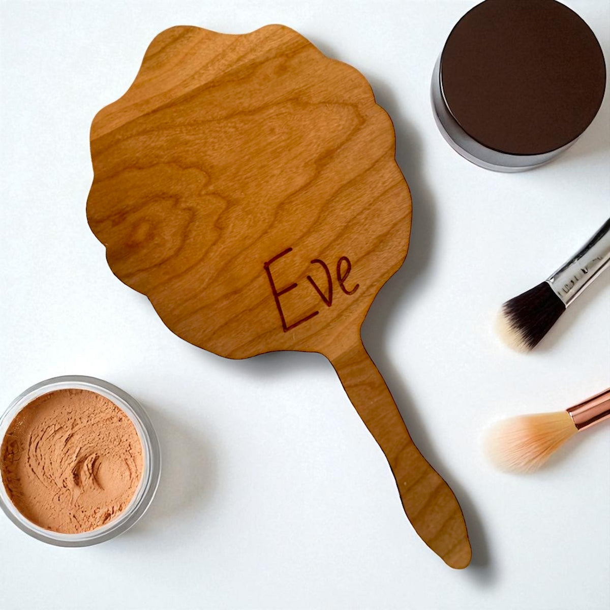 Personalized scalloped wooden hand mirror with the name "Eve" engraved, surrounded by makeup.
