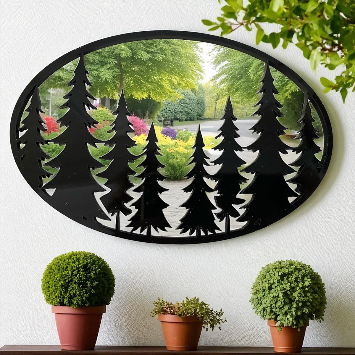 Oval black garden mirror with a pine tree silhouette overlay, mounted on a white wall above three potted plants.
