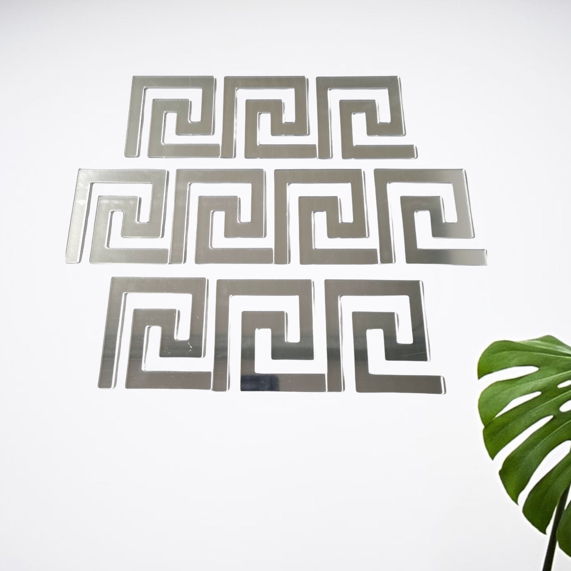 Silver mirror-finish geometric Greek key tiles arranged on a white wall with a green plant leaf in the corner.