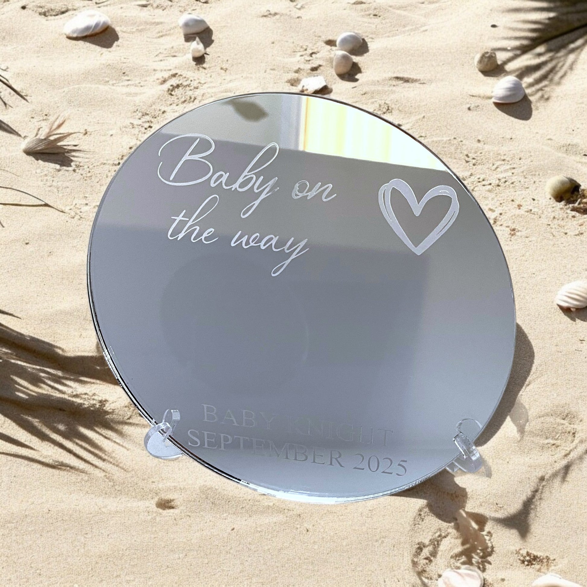 Round personalized mirror on sand, engraved with "Baby on the way" and custom text.