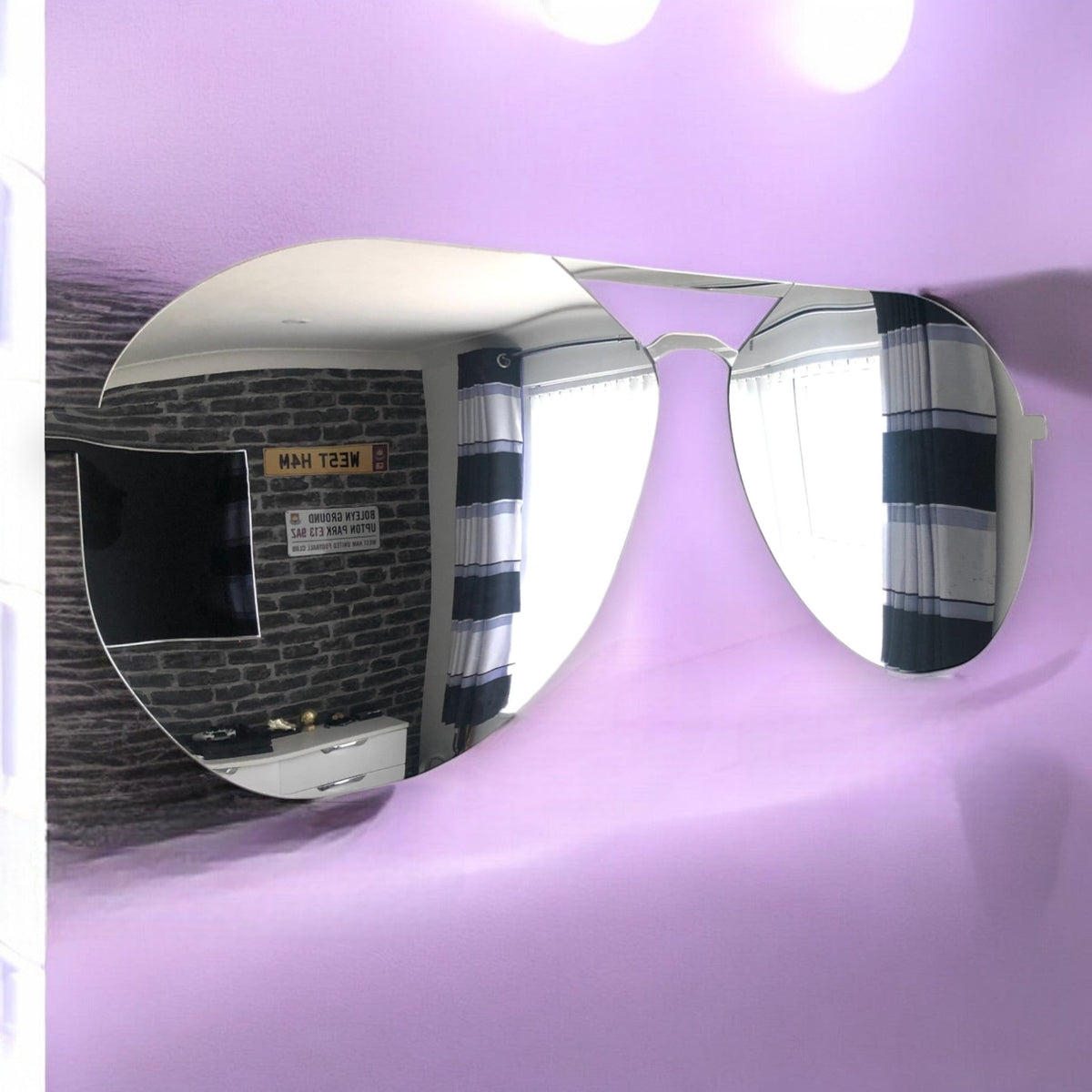 Aviator sunglasses-shaped wall mirror mounted on a light purple wall, reflecting a room.