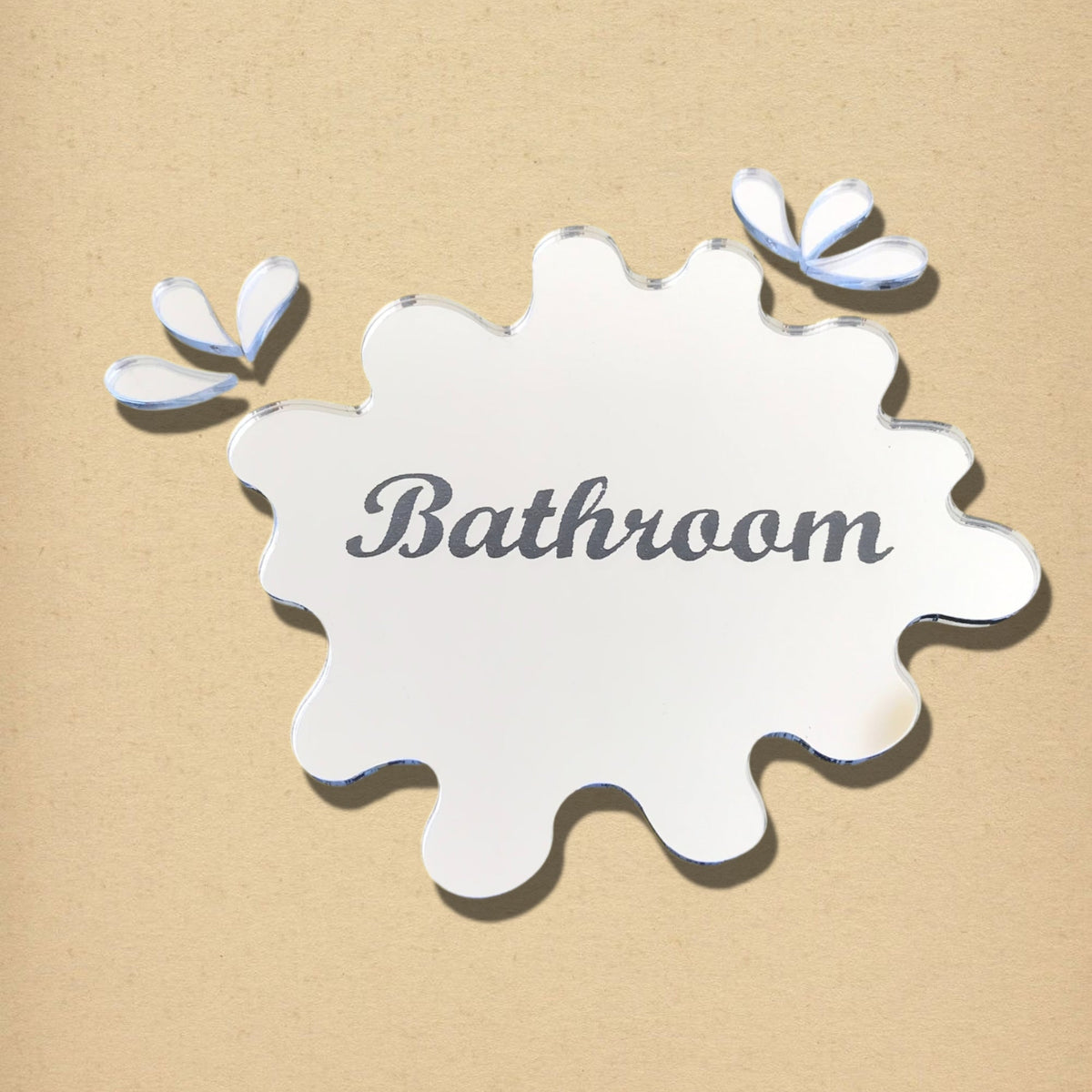 Mirrored puddle-shaped bathroom sign with engraved text and two separate droplet pieces on beige background.