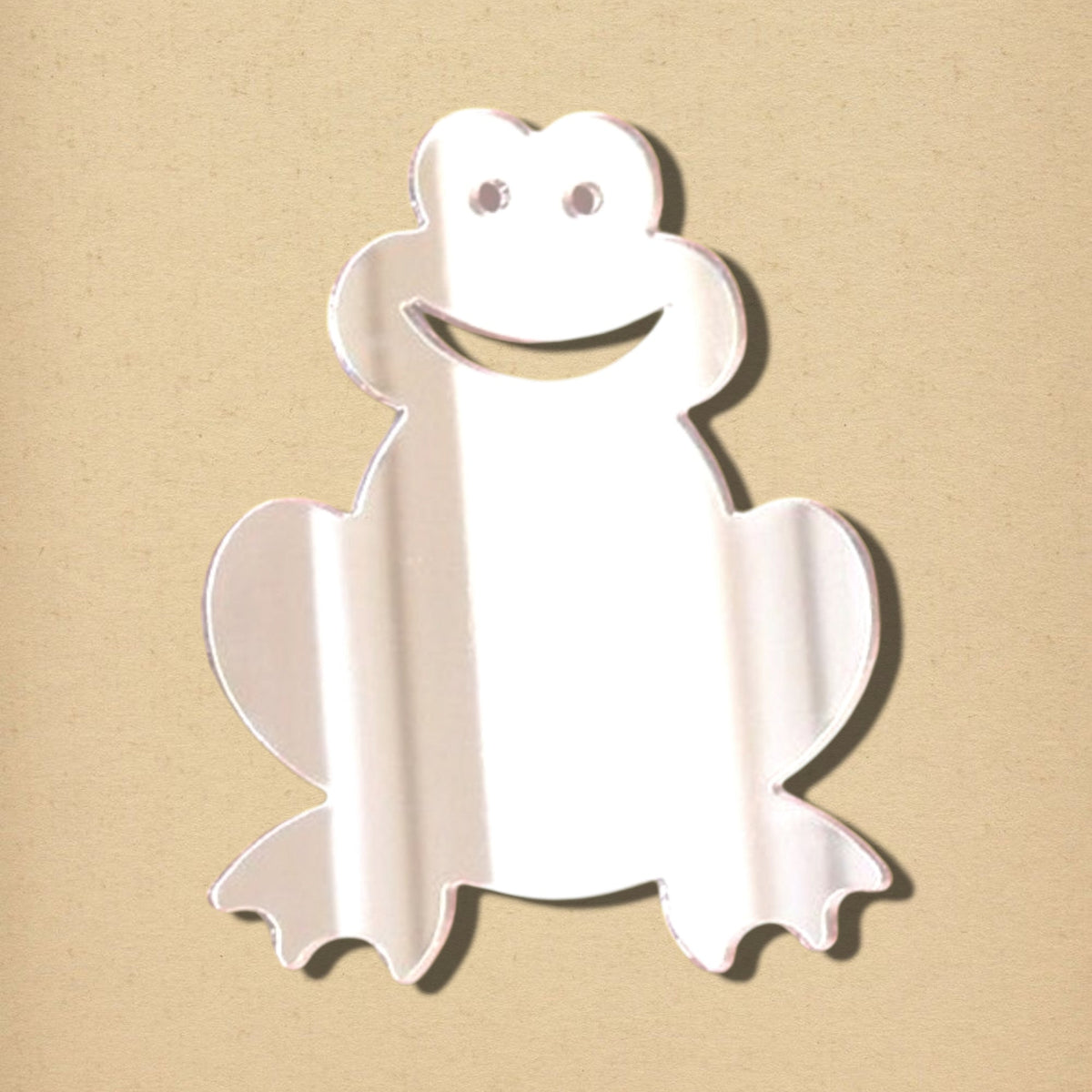 Frog-shaped mirror made of reflective material with cutouts for eyes and a smiling mouth, set against a plain beige background.