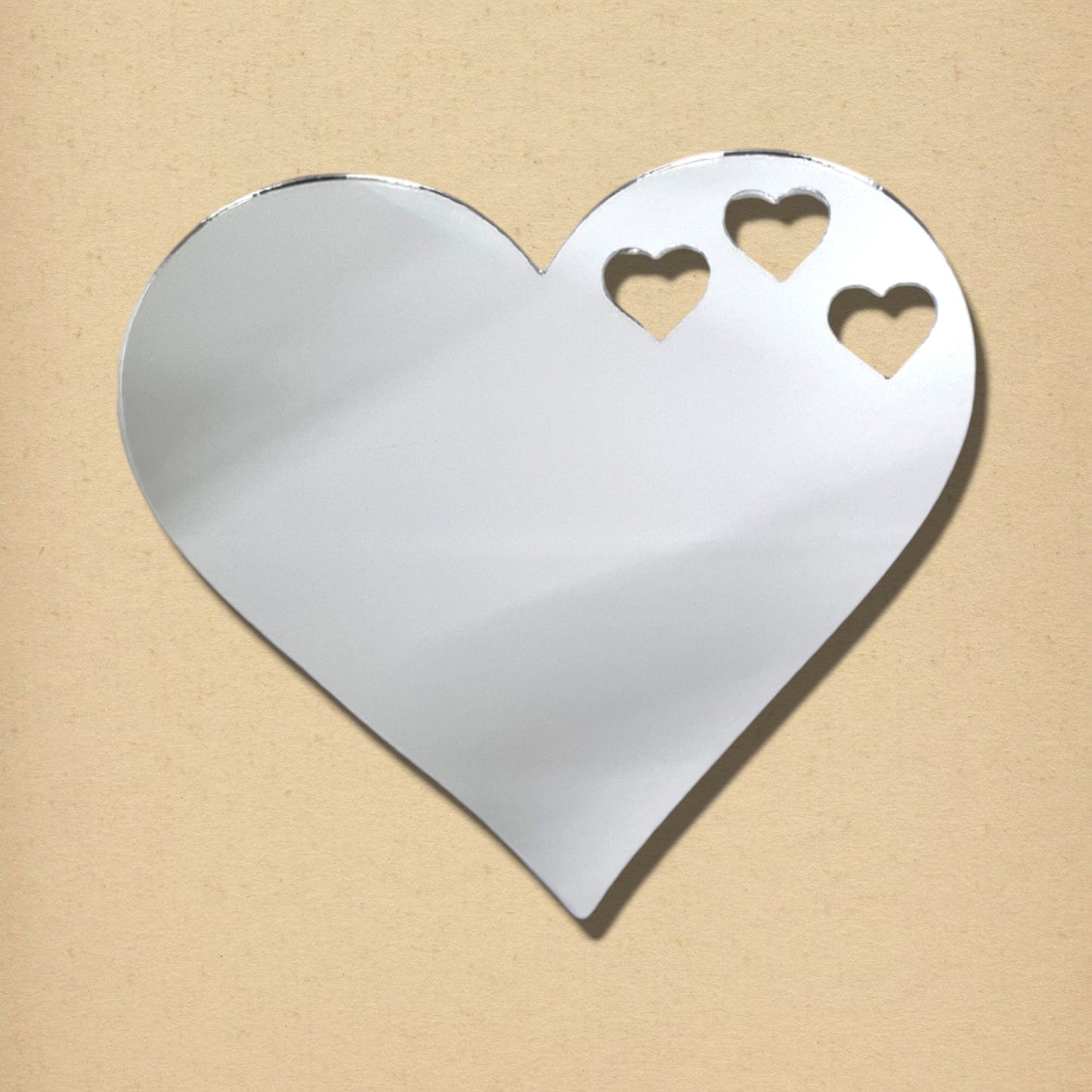 Silver heart-shaped acrylic mirror with three small heart cutouts near the top right, on a tan background.