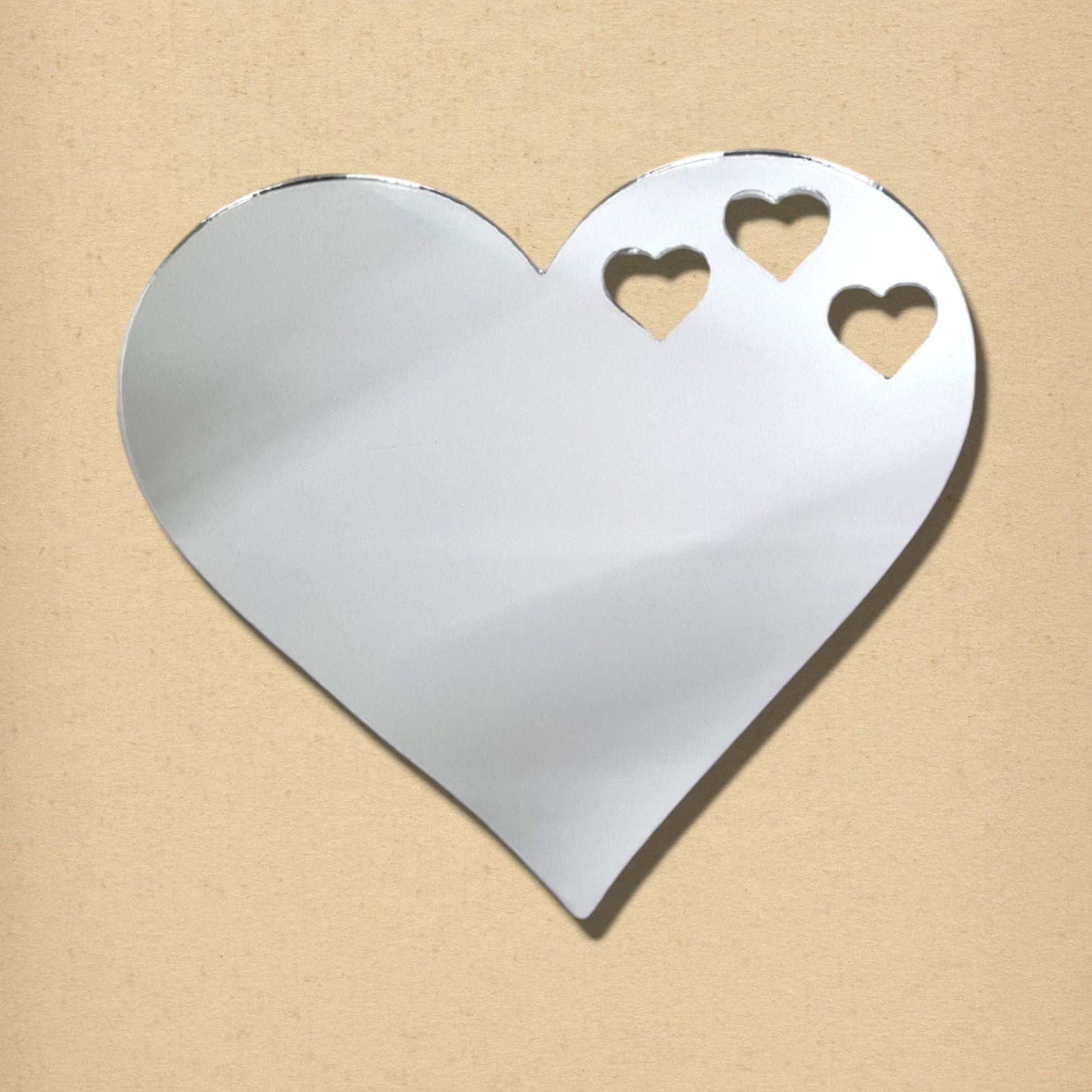 Silver heart-shaped acrylic mirror with three small heart cutouts near the top right, on a tan background.