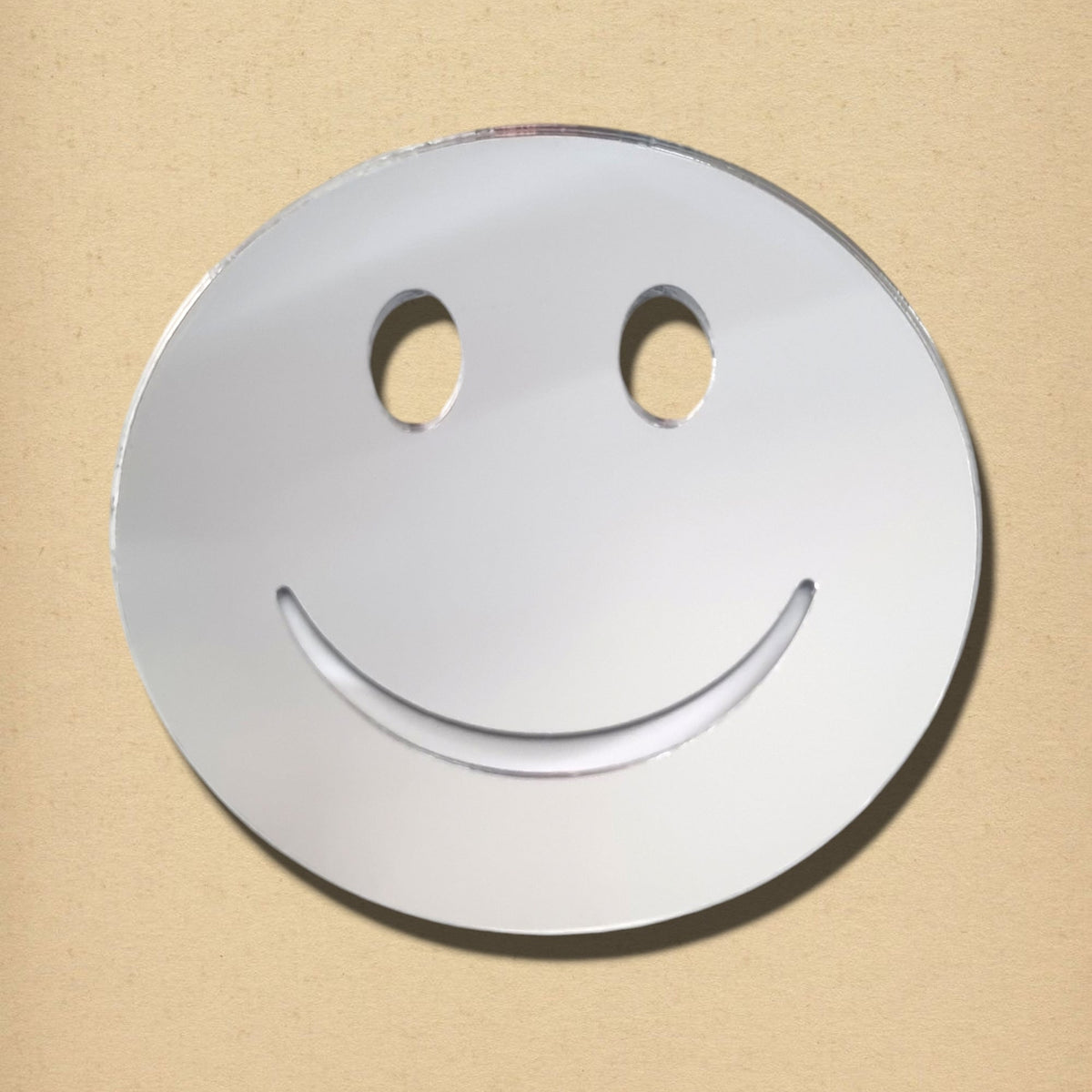 A round, silver smiley face mirror with oval eye cutouts and an upturned smile cut against a beige background.